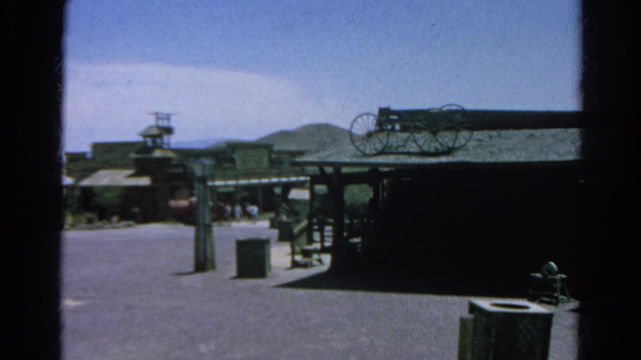 1966 Silver Lake, California home-movie frame showing a Western-themed street with wooden facades, wagon wheels, hills, and distant visitors.