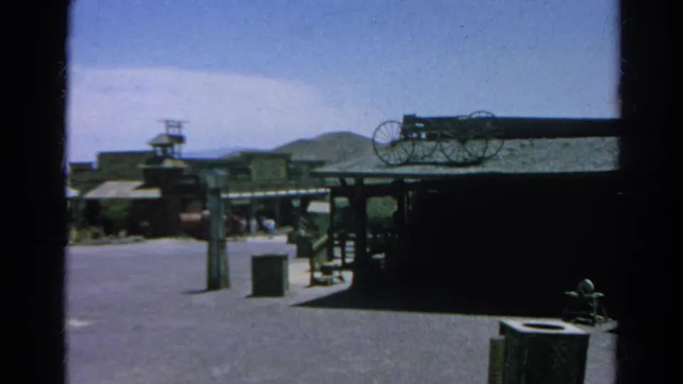 1966 Silver Lake, California home-movie frame showing a Western-themed street with wooden facades, wagon wheels, hills, and distant visitors.