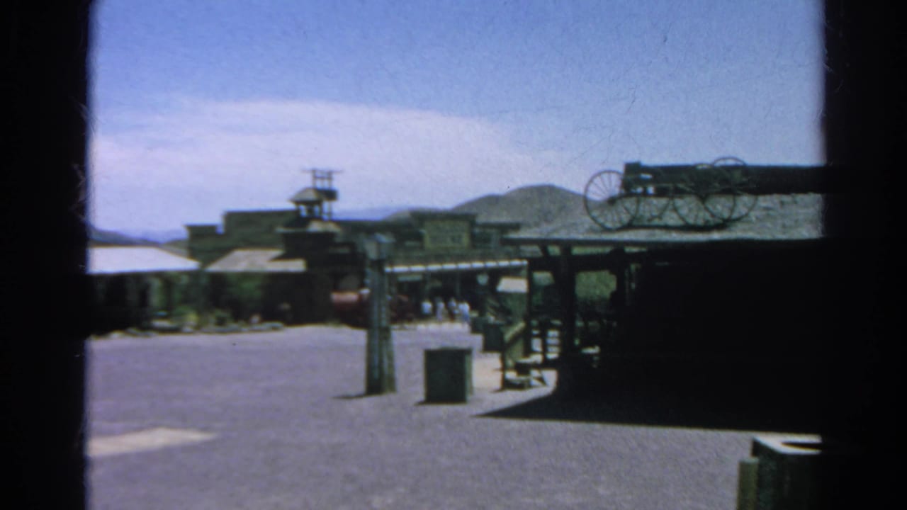 Wide street view with Western facades, wagon-wheel display, shade structure, hills, and distant people.