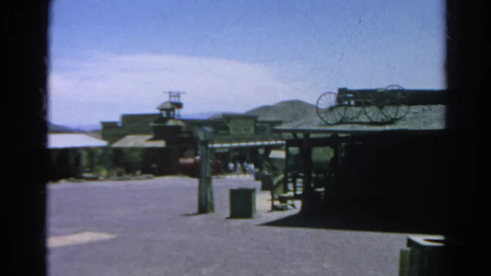 1966 Silver Lake, California frame with Western-style storefronts, a wagon-wheel display, open pavement, hills, and small distant visitors.