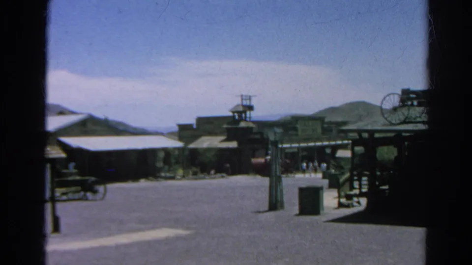 1966 Silver Lake, California home-movie view of a Western-style street, low buildings, wagon-wheel detail, distant visitors, and hills.