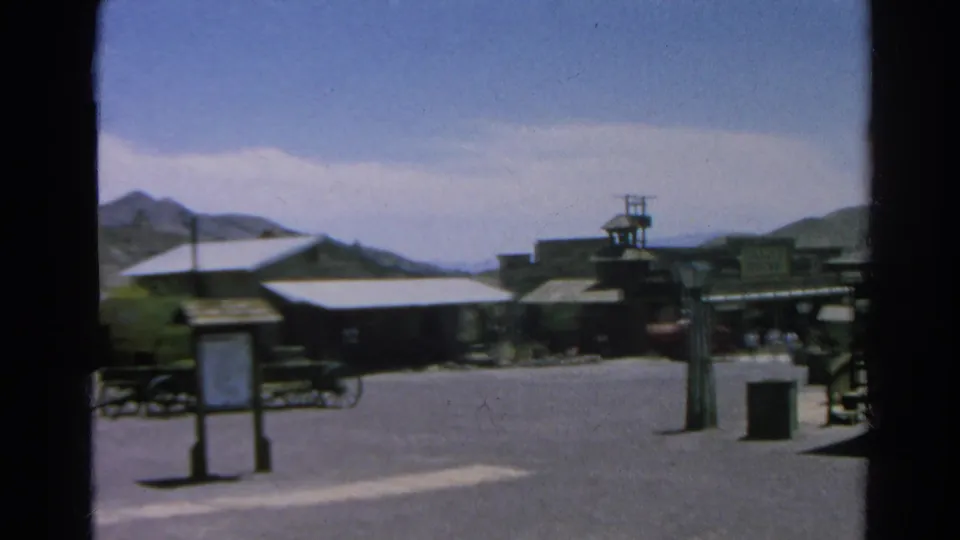 1966 Silver Lake, California frame showing a signboard, Western-style buildings, wagon-wheel display, open street, hills, and distant visitors.