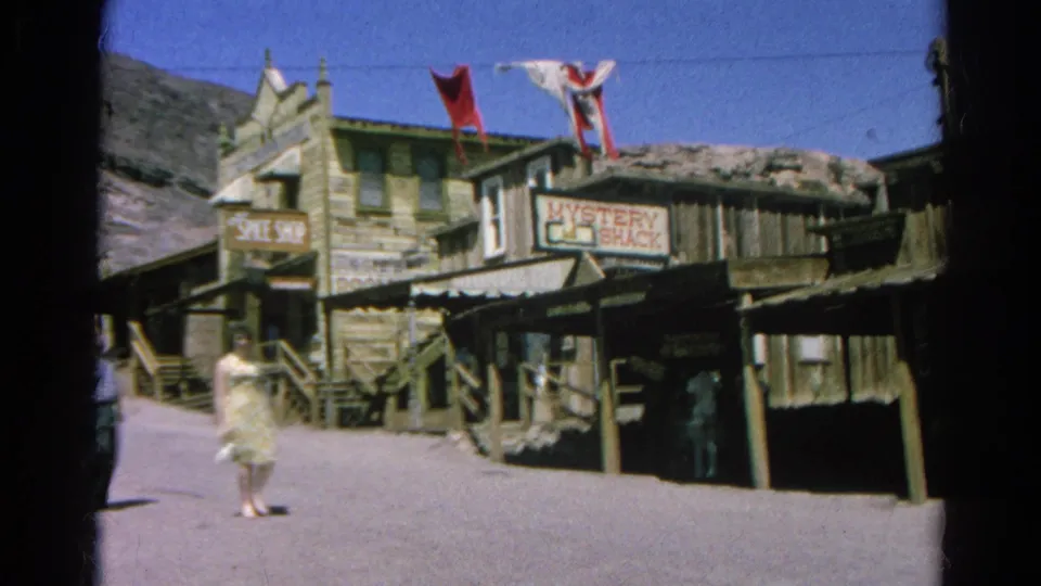 A woman in a light 1960s dress walks through a Silver Lake western-themed street with wooden shops and signs behind her.