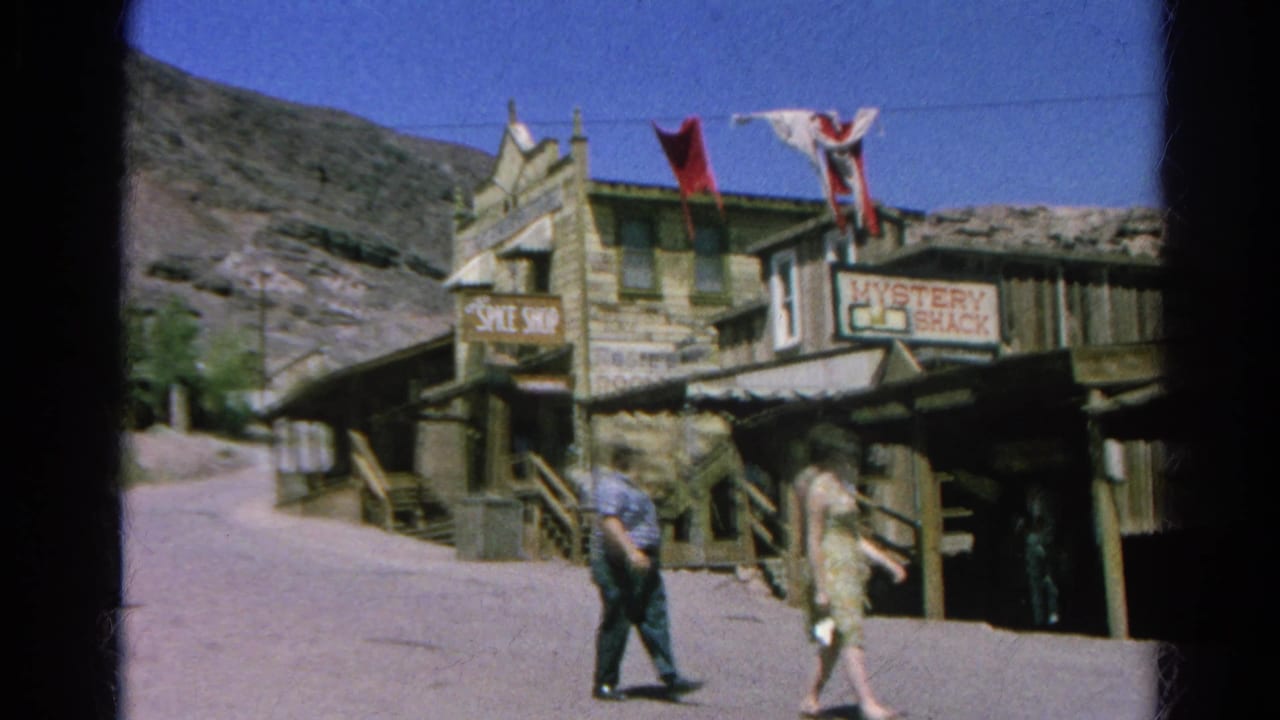 Two adults walk along a sunlit street beside rustic wooden buildings and hanging flags.