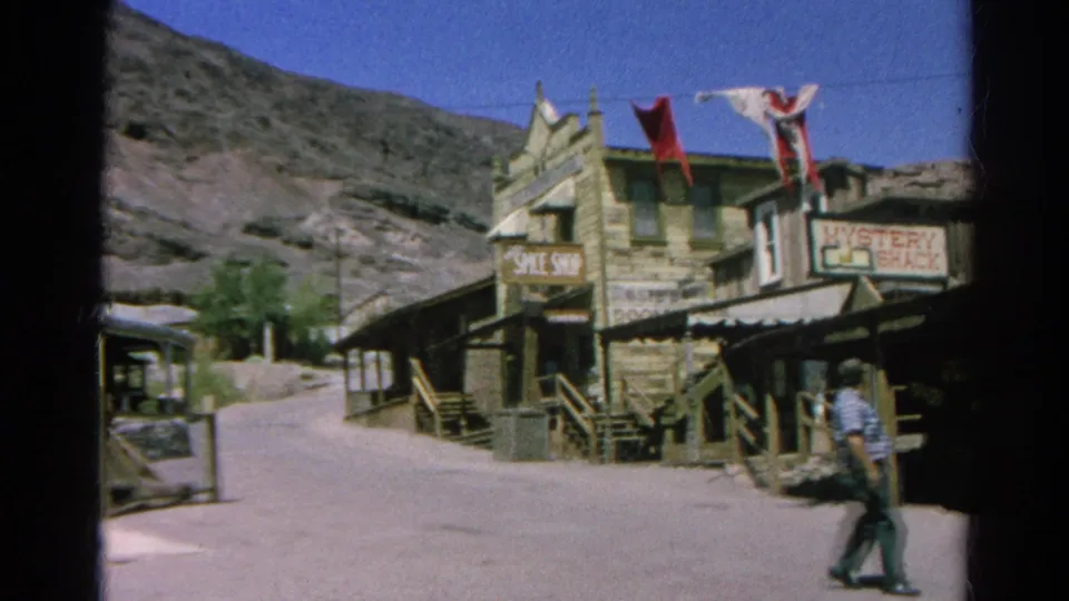 1966 8mm view of a man walking past a Western-style street scene with signs, stairs, railings and dry hills.