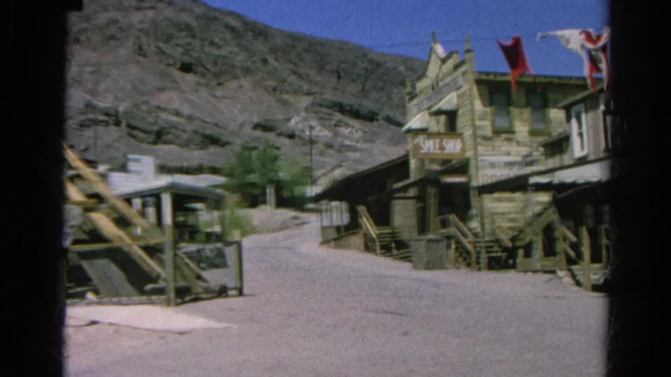 Silver Lake 1966 frame showing the Western-style storefront row after the walkers pass, with flags and dry hillside visible.