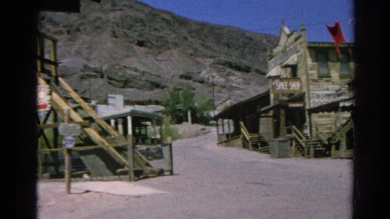 1966 8mm view of a quiet Silver Lake, California village street with wooden buildings, signage, a red flag, and dry hills behind.