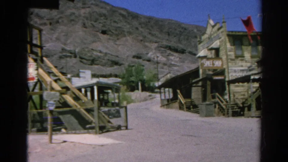 1966 8mm view of a quiet Silver Lake, California village street with wooden buildings, signage, a red flag, and dry hills behind.