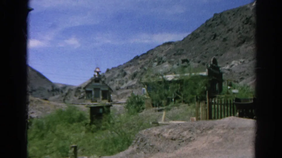 1966 Silver Lake home-movie frame with a small towered building, hillside backdrop, shrubs, fence posts, and an empty foreground mound.