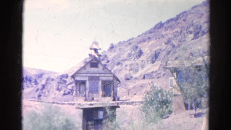 1966 8mm view of a weathered wooden building with a small bell tower, scrub vegetation, and rocky hills in Silver Lake, California.