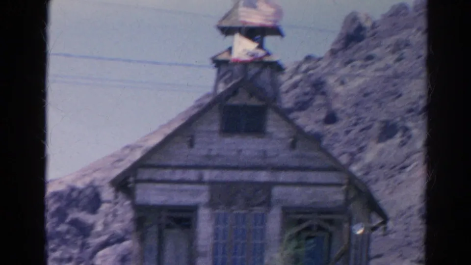 Closer 8mm frame of the Silver Lake wooden building, showing the roofline, tower, American flag, and power lines against the hillside.