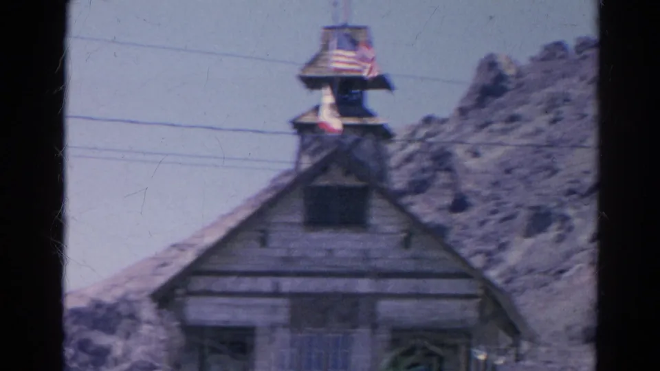 Close 1966 home-movie view of the weathered facade and bell tower, with an American flag above and rocky Silver Lake hills behind.