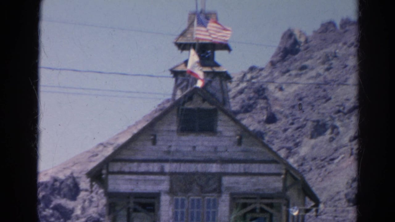 1966 Silver Lake 8mm frame showing a rustic gabled wooden building, small tower, American flag, and rocky hillside behind it.