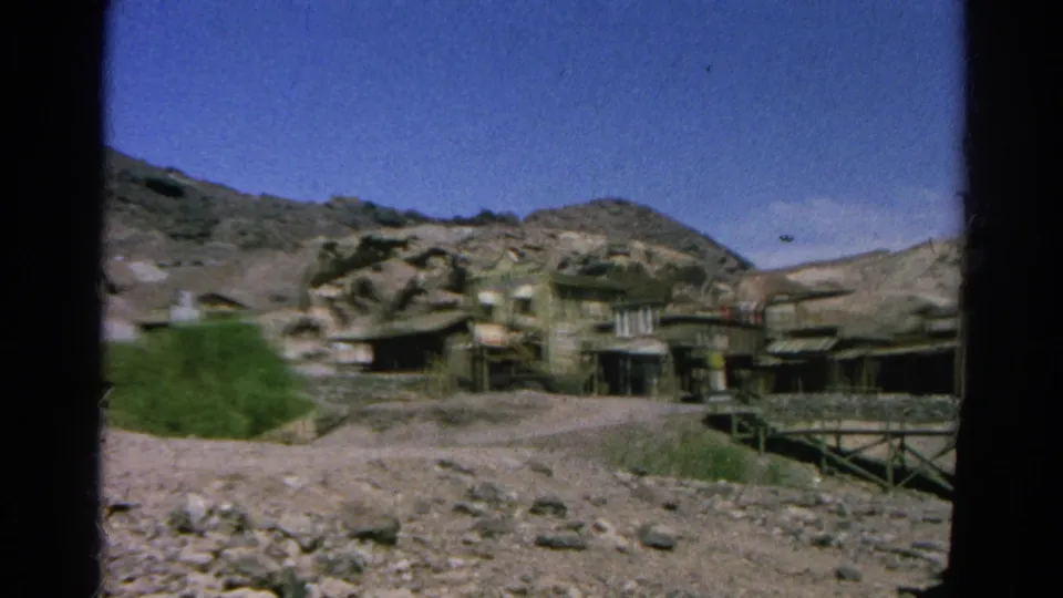 1966 Silver Lake 8mm wide shot showing a sparse settlement of wooden buildings, scrub vegetation, dusty ground, and rocky hills.