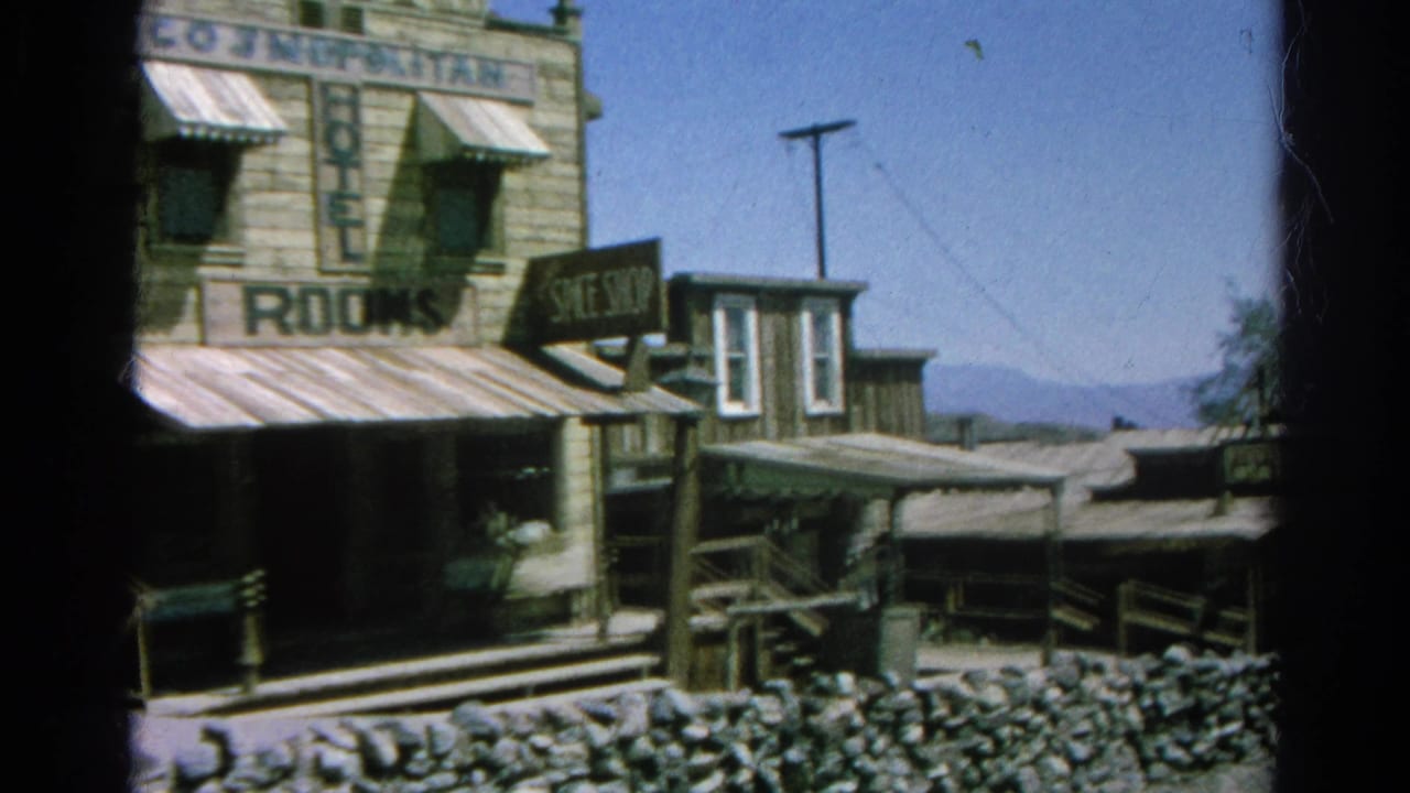Western-style wooden buildings with rooms and shoe shop signs beside a stone wall.