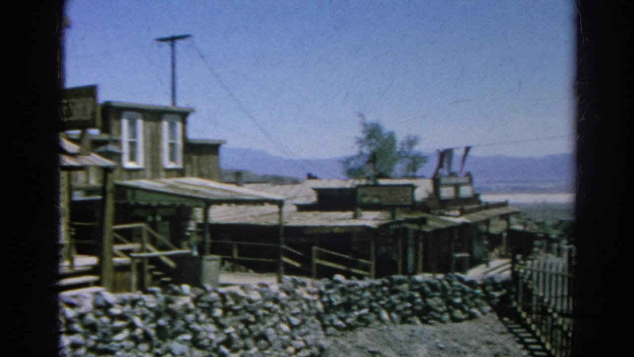 The pan continues across weathered Western-style buildings, porch railings, a stone wall, and mountains beyond the town set.