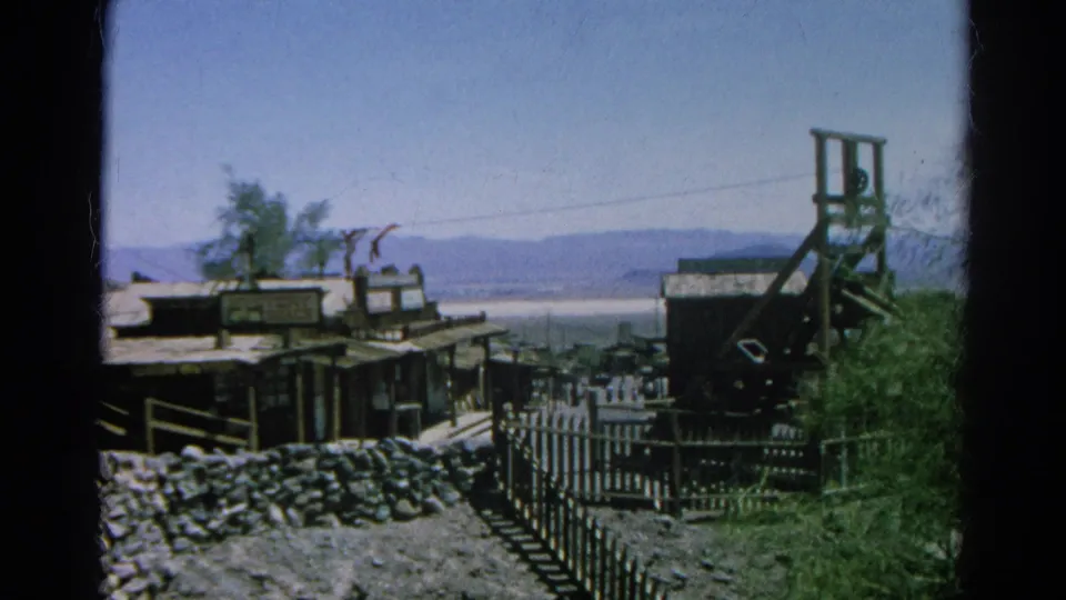 Final frame shows the Western-style buildings, fence, stone wall, distant lake and mountains, with a tall wooden tower at right.