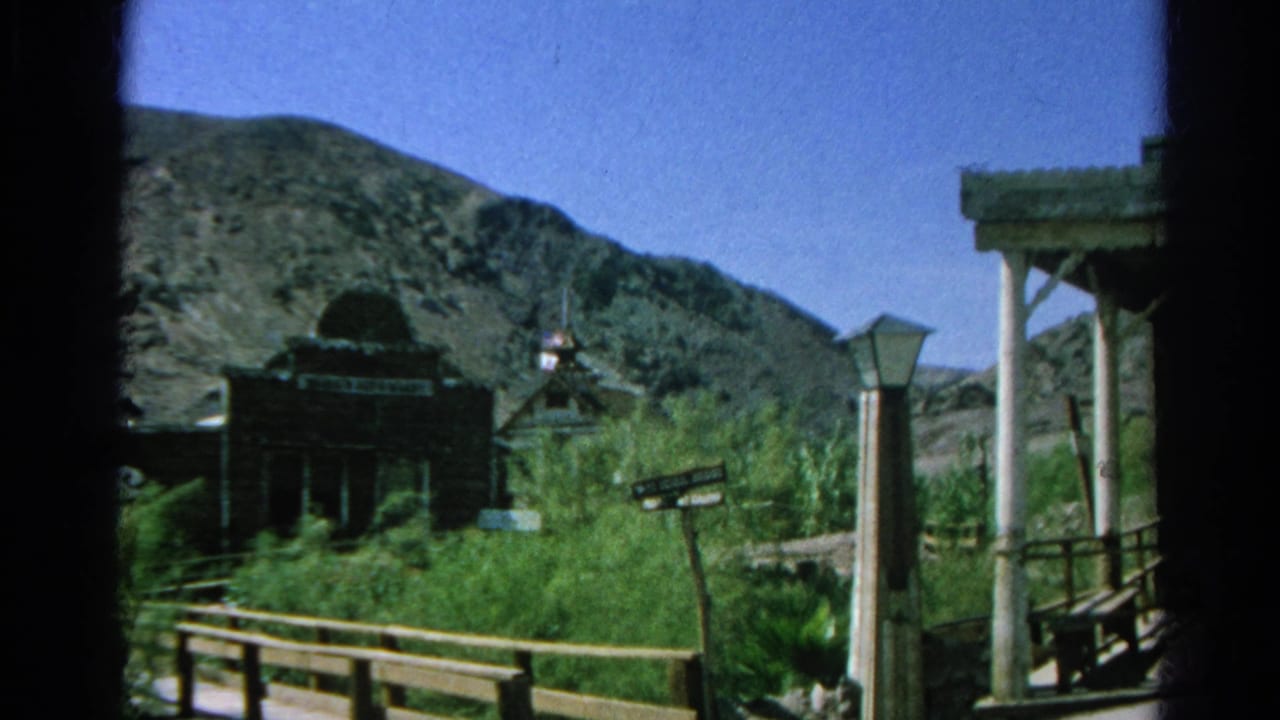 Dark wooden church-like building, brush, signpost, porch posts, and rocky hillside.