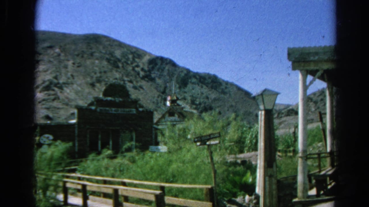 Western-style storefronts, fence rails, green shrubs, and rocky hills under a blue sky.