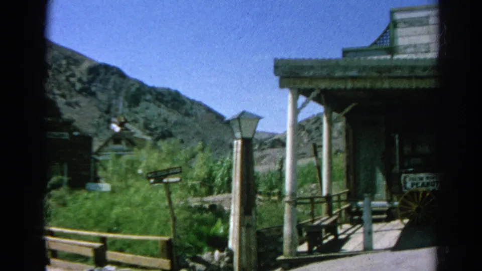 1966 Silver Lake 8mm frame showing a Western porch, fence rail, lamppost, shrubs, hills, and storefront signage.