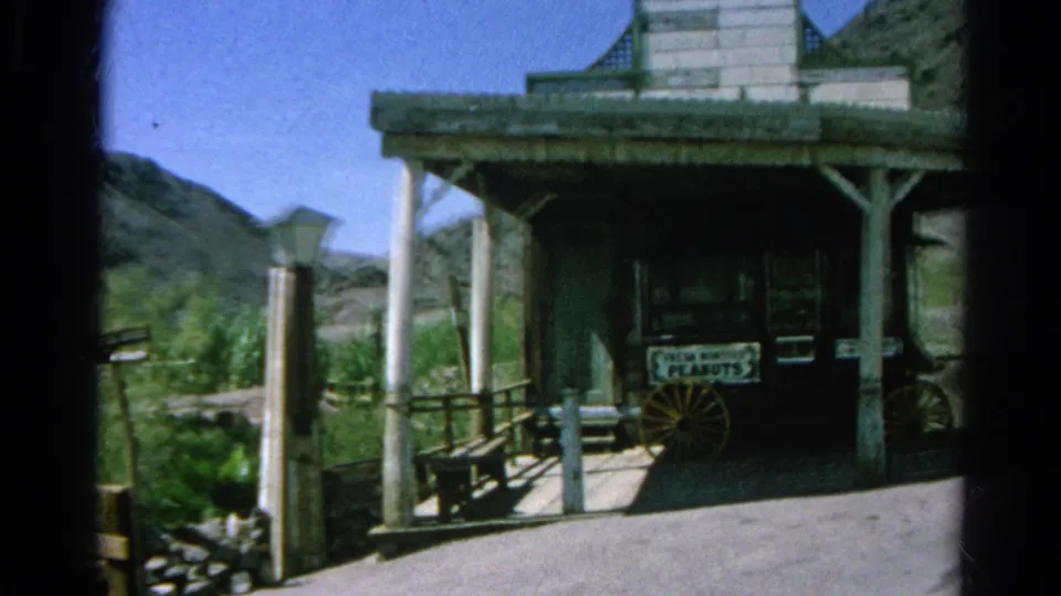 1966 Silver Lake frame centered on a Western-style porch with a Fresh Roasted Peanuts sign, wagon wheel, lamppost, shrubs, and hills.