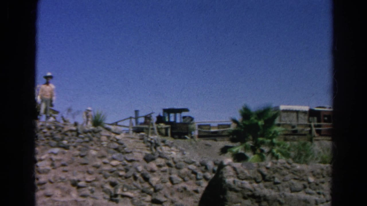 Rocky hillside with a man, wooden fencing, small rail car, and palm tree in a dry landscape.