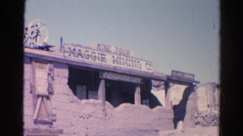 1966 8mm frame of the Maggie Mining Company mine-tour facade, with commissary signage, mining equipment, and pale desert sky.