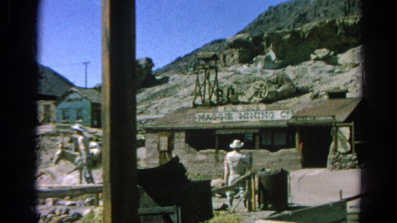 Western mining-town facade with Maggie Mining Co. sign, rocky hills, and adults in cowboy hats.