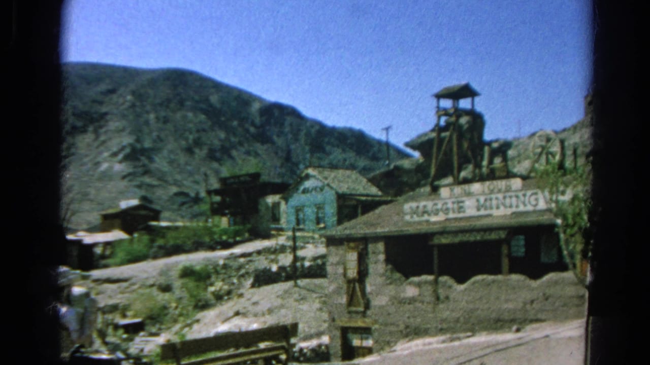 End of the 1966 pan on the Maggie Mining Co. building, mine tower, arid hills, and dusty road with partial visitors at the edge.