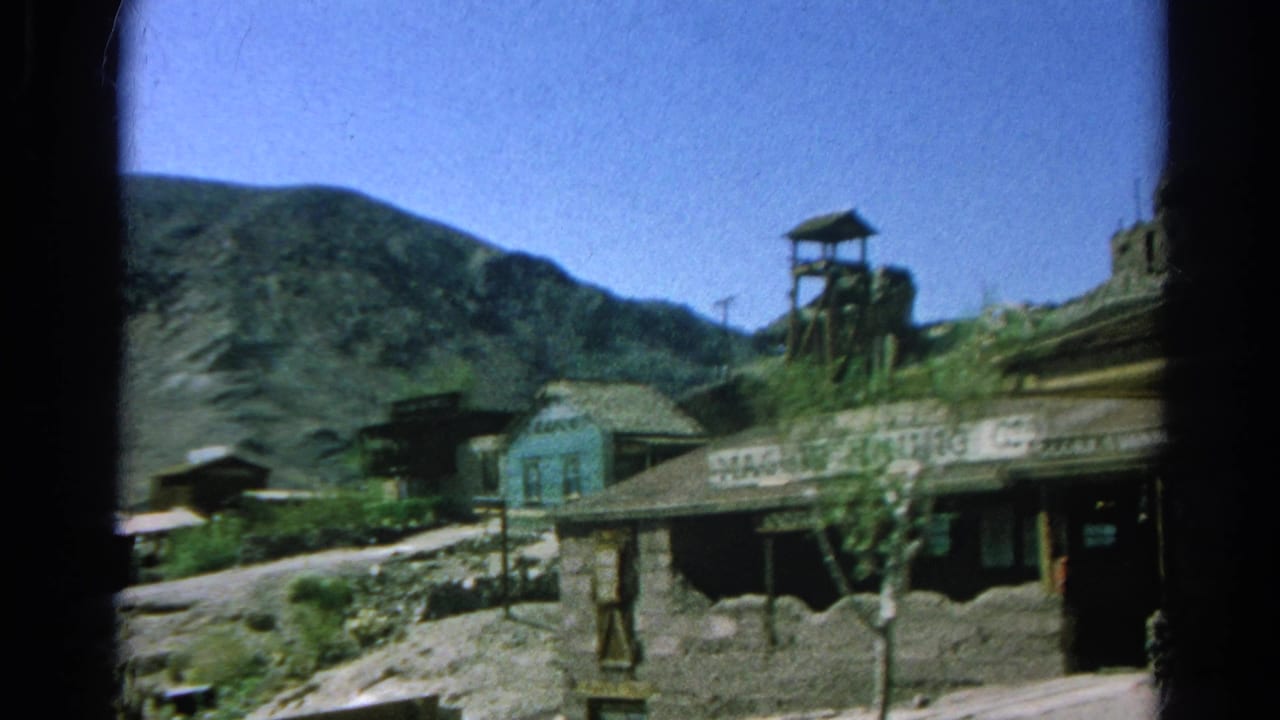 Weathered wooden mining-town buildings with mountain slopes, a headframe, and MAGGIE MINING CO. signage.