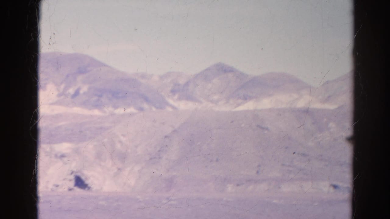The 1966 home-movie frame shows low desert hills and sandy foreground through a dark-framed window opening.
