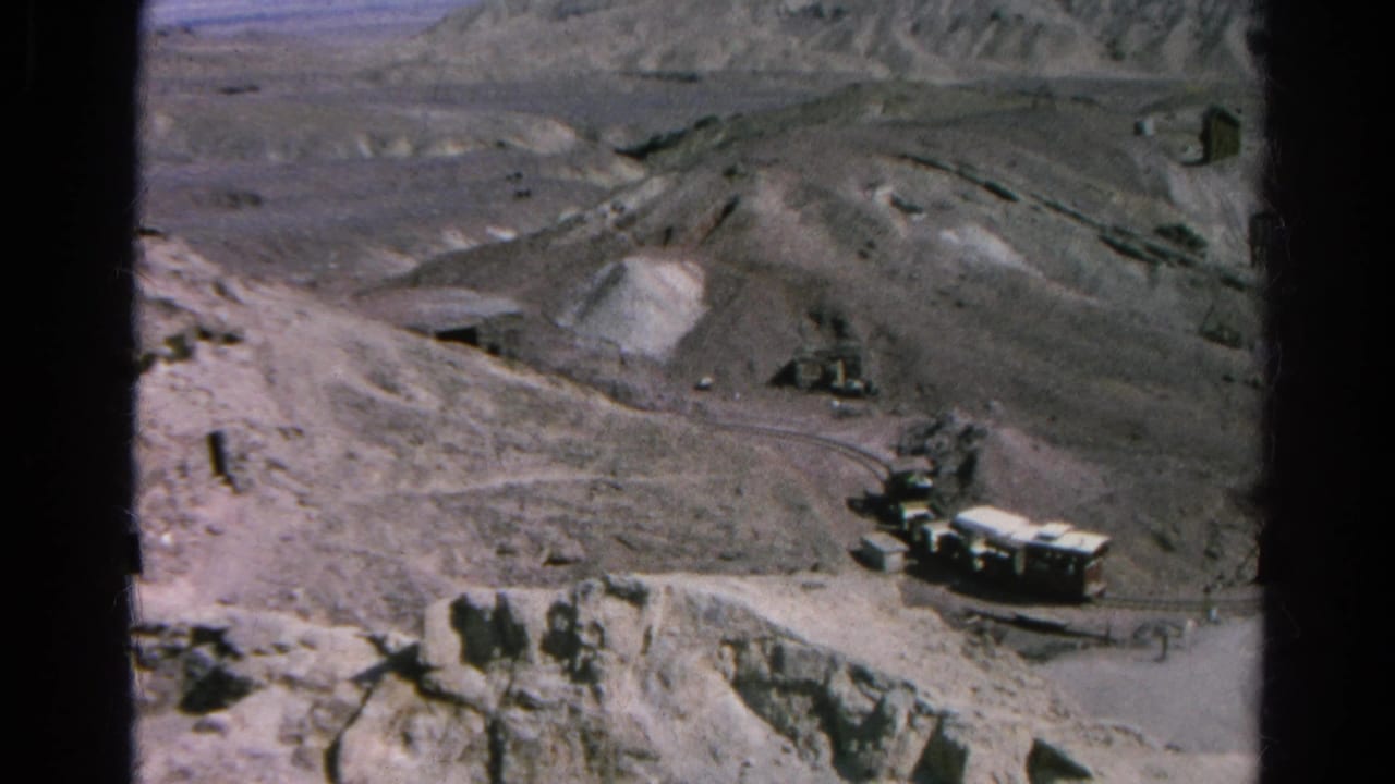 Trucks and pale railcars or trailers sit below excavated hillsides.