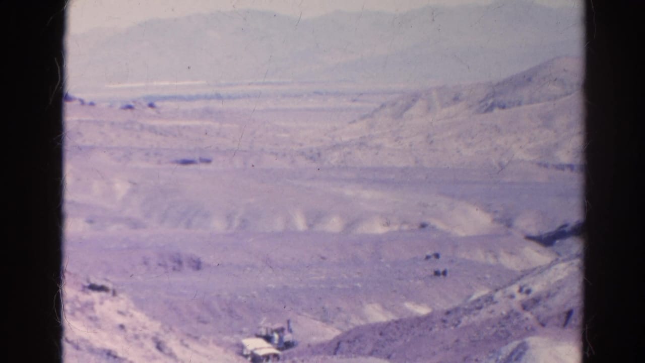 Wide 1966 Silver Lake desert frame with pale hills, a road corridor, distant mountains, and small indistinct objects in the valley.