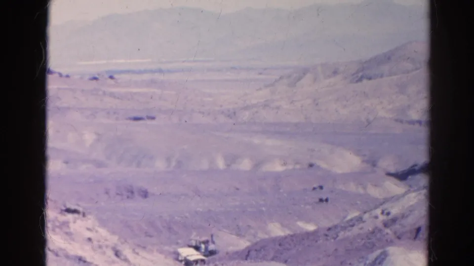 Wide 1966 Silver Lake desert frame with pale hills, a road corridor, distant mountains, and small indistinct objects in the valley.
