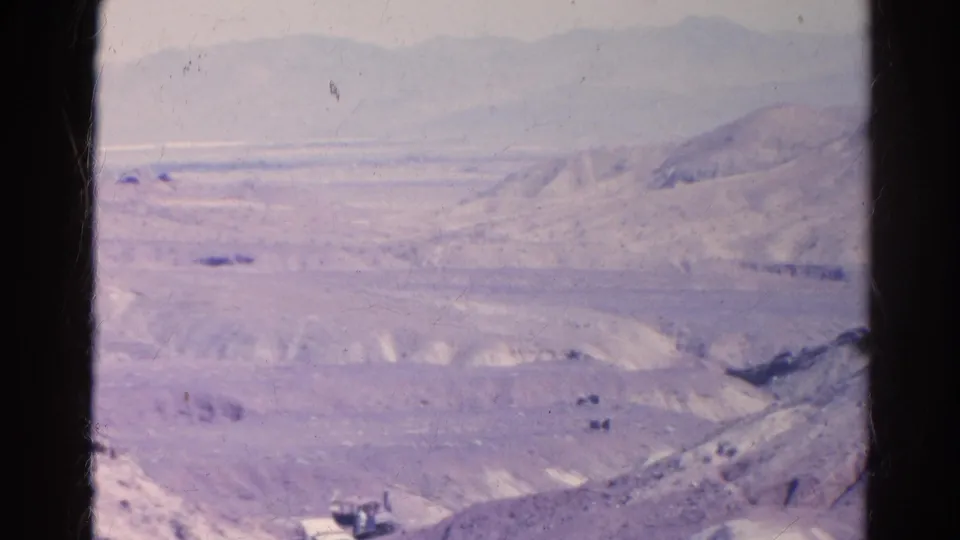 1966 8mm view near Silver Lake, California, showing pale desert ridges, hazy mountains, and a small train or rail vehicle at the bottom of frame.