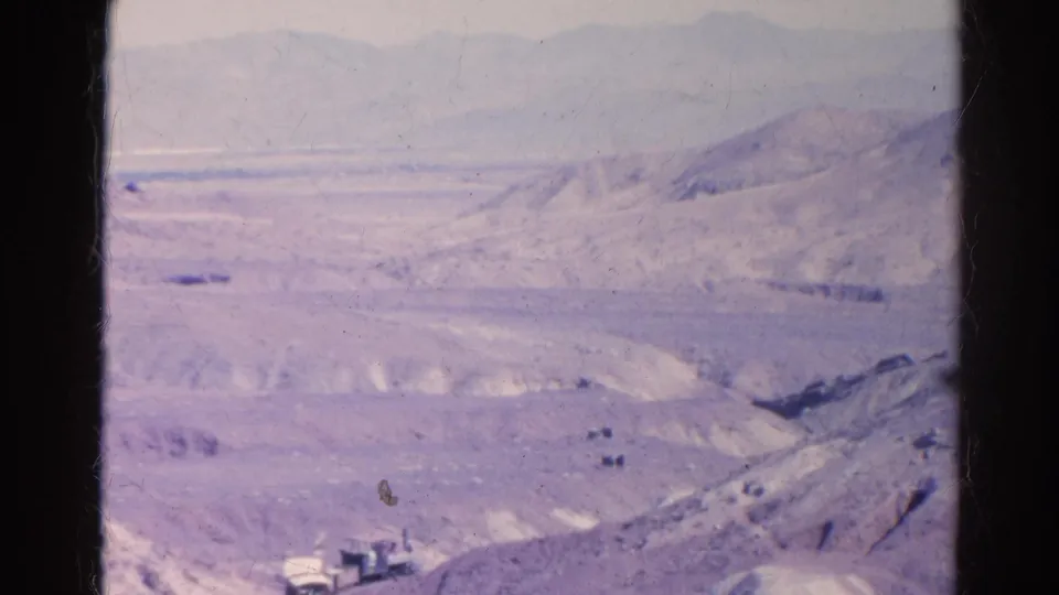 Wide 8mm desert view near Silver Lake, California, with a small train or rail vehicle crossing the lower frame beneath hazy mountain ridges.
