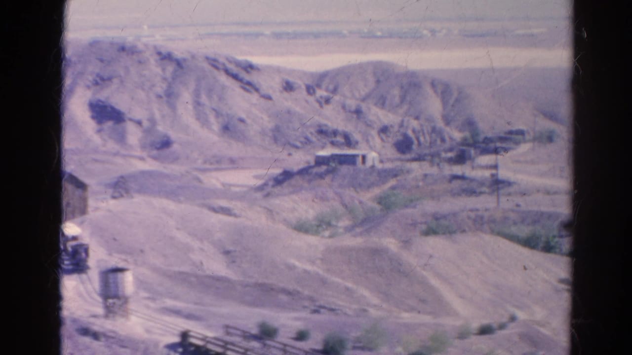 Small train and water tower in a dry hilly landscape near railroad tracks.