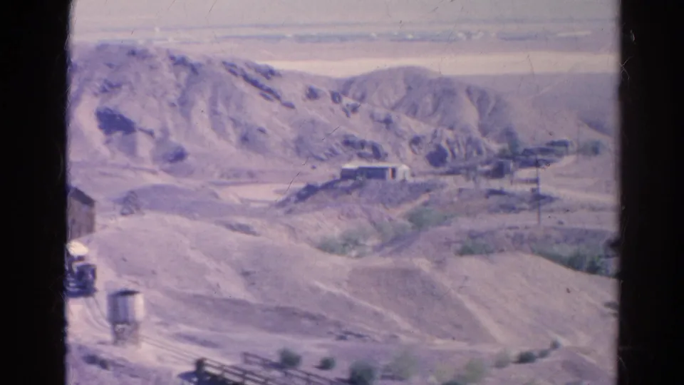 1966 8mm frame from Silver Lake, California, showing a distant small train area, water tower, dry hills, and sparse desert vegetation.