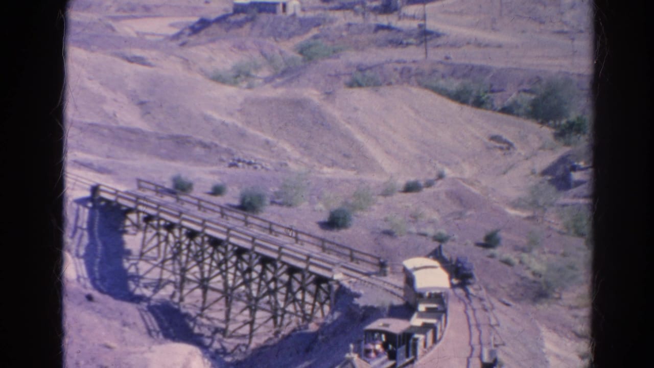 1966 home-movie frame of the small train exiting the trestle, with open cars following over the dry hillside track.