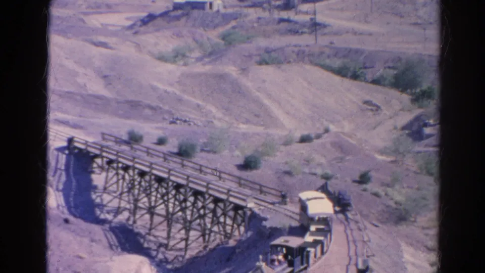 1966 home-movie frame of the small train exiting the trestle, with open cars following over the dry hillside track.