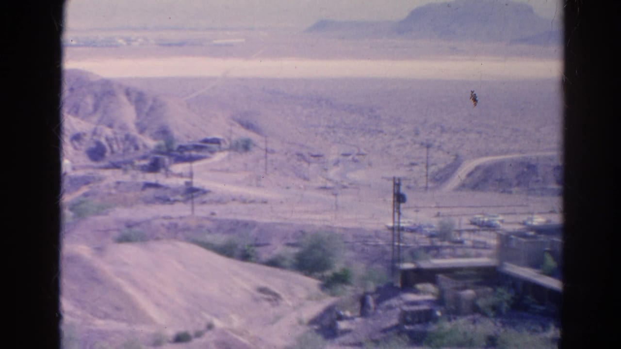 Distant desert settlement with dirt road, utility poles, tower, and hazy hills.