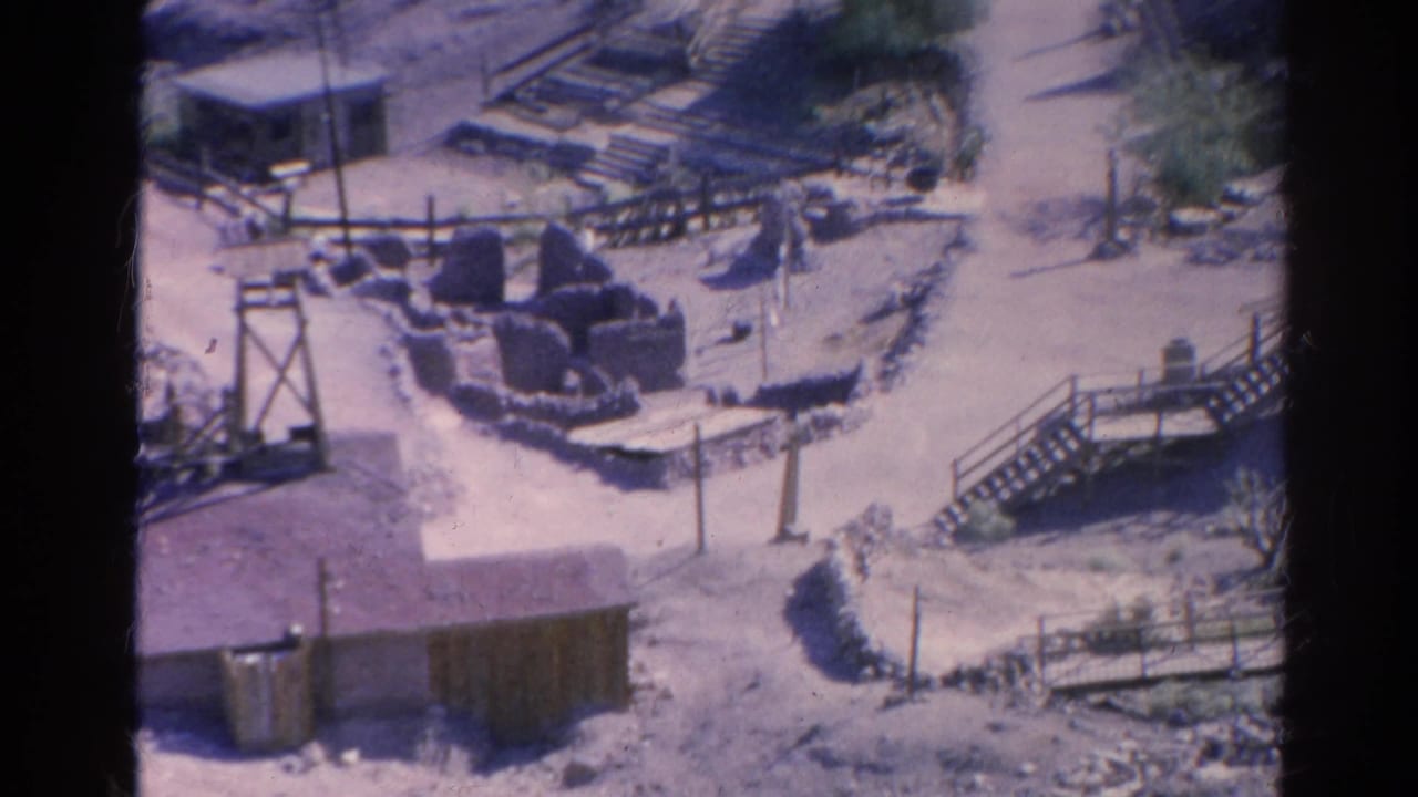1966 8mm frame of a desert settlement area with wooden stairs, railings, low structures, sandy paths, and sparse vegetation.