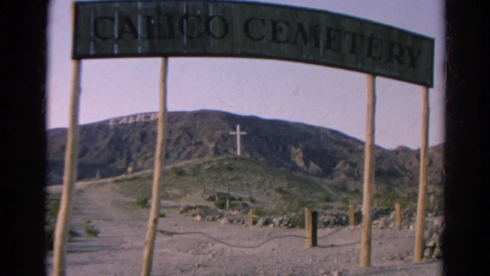 Calico Cemetery entrance appears in 1966 California 8mm footage, with a dirt road, hillside lettering, and a white cross beyond the sign.