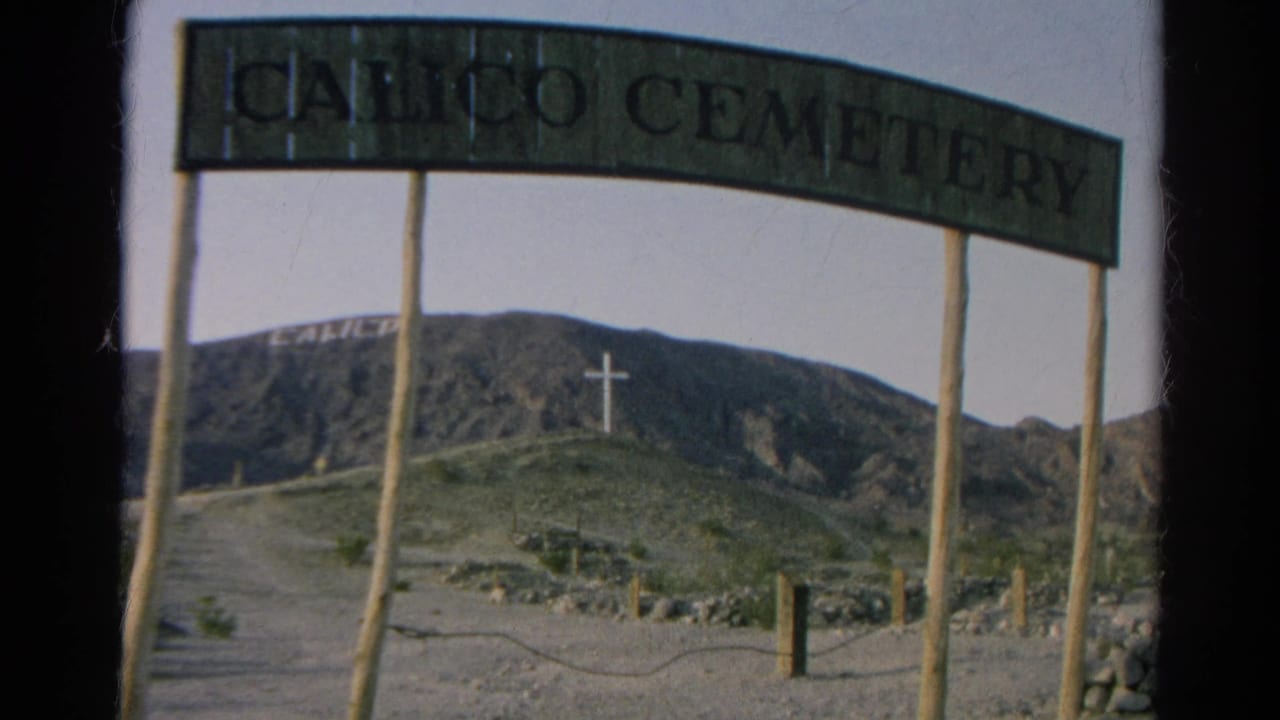 1966 8mm frame of the Calico Cemetery entrance sign with wooden posts, a dirt path, rocky hillside, and white cross in the distance.