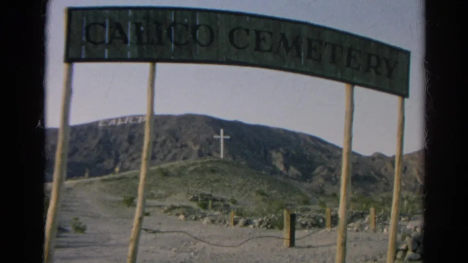 1966 8mm frame of the Calico Cemetery entrance sign with wooden posts, a dirt path, rocky hillside, and white cross in the distance.