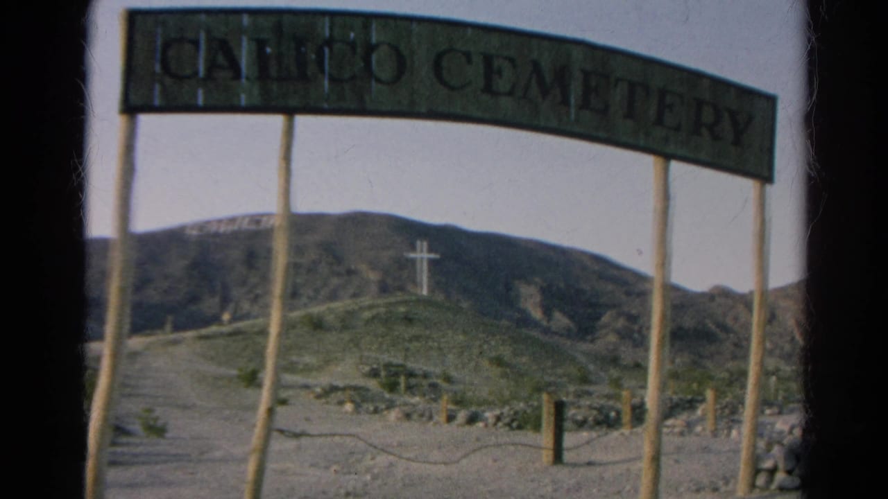 Visitor-shot 1966 view of the Calico Cemetery sign, desert path, hillside cross, rocky terrain, and distant mountain backdrop.