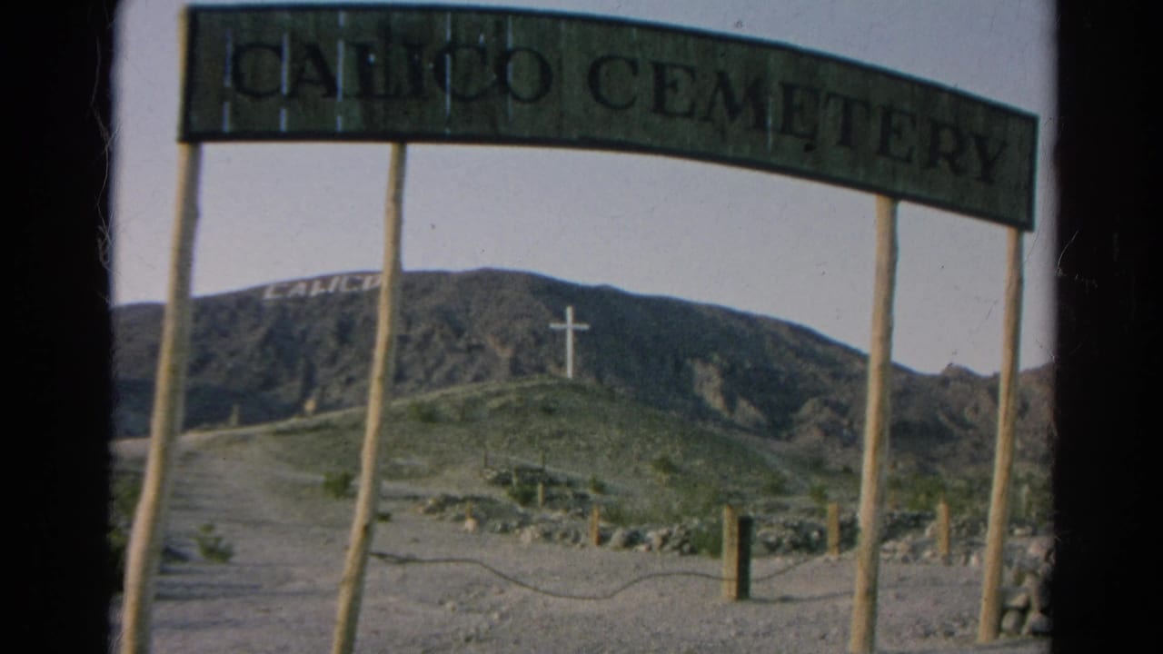 1966 8mm frame showing the Calico Cemetery sign cropped close, with a dirt approach, wooden posts, mountain lettering, and hilltop cross.