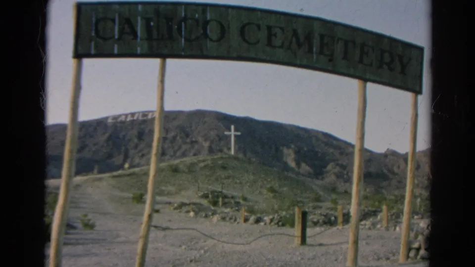 1966 8mm frame showing the Calico Cemetery sign cropped close, with a dirt approach, wooden posts, mountain lettering, and hilltop cross.