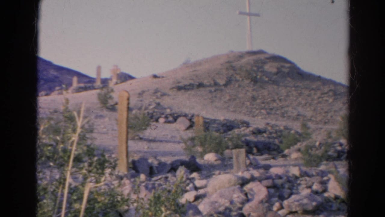 1966 8mm view inside or near Calico Cemetery, showing rocky gravesite terrain, wooden markers, sparse desert plants, and a white cross on the hill.