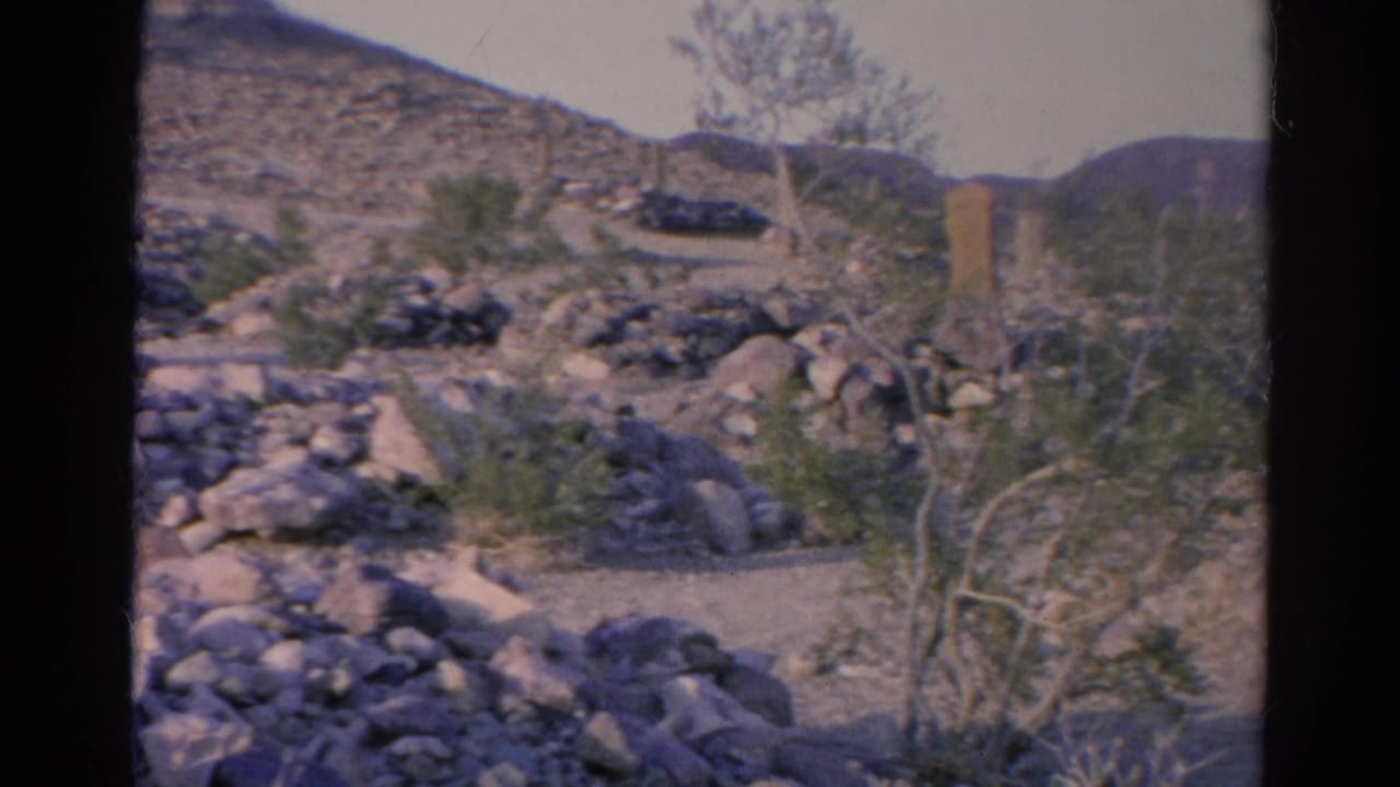 1966 home-movie frame of a dirt path through rocky Silver Lake terrain, with sparse desert plants and marker-like wooden posts nearby.